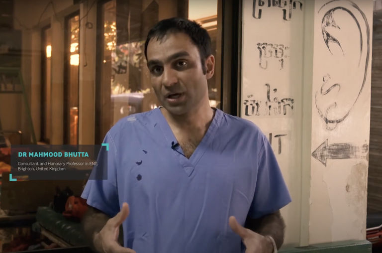 Person in blue medical scrubs standing outside building with caption identifying Dr Mahmood Bhutta, ENT Consultant in Brighton, UK