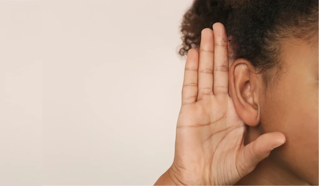 Person holding hand to ear to emphasise active listening and hearing health, close-up of ear and hand gesture on light background.