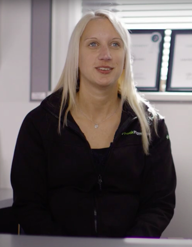 Individual wearing Peak Pharmacy jacket seated indoors with framed certificates on the wall