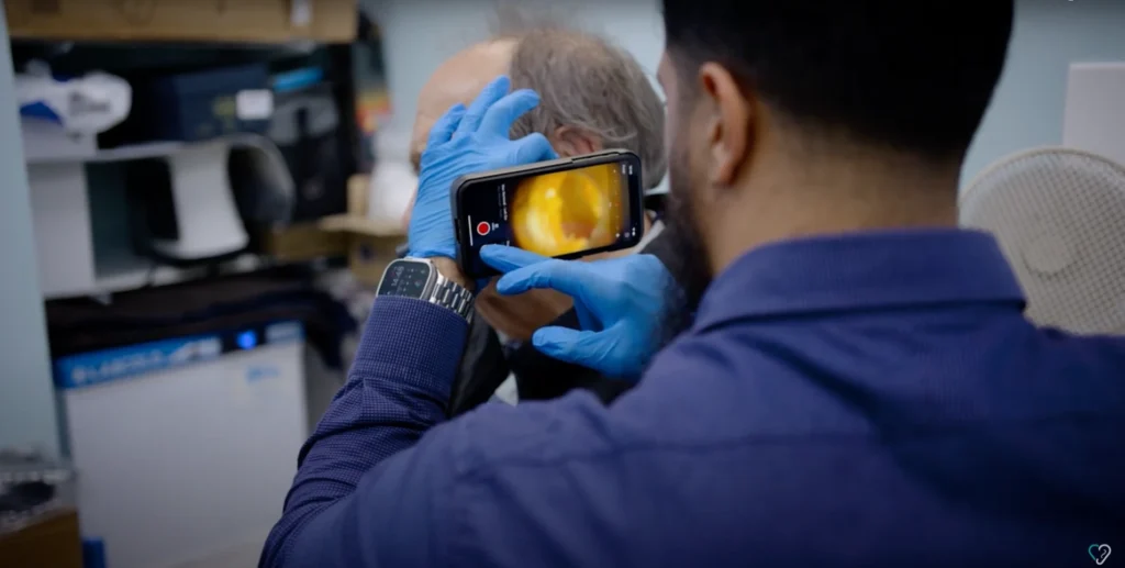 Person wearing blue gloves using Tympa to examine ear with magnified view on screen, in medical or office setting
